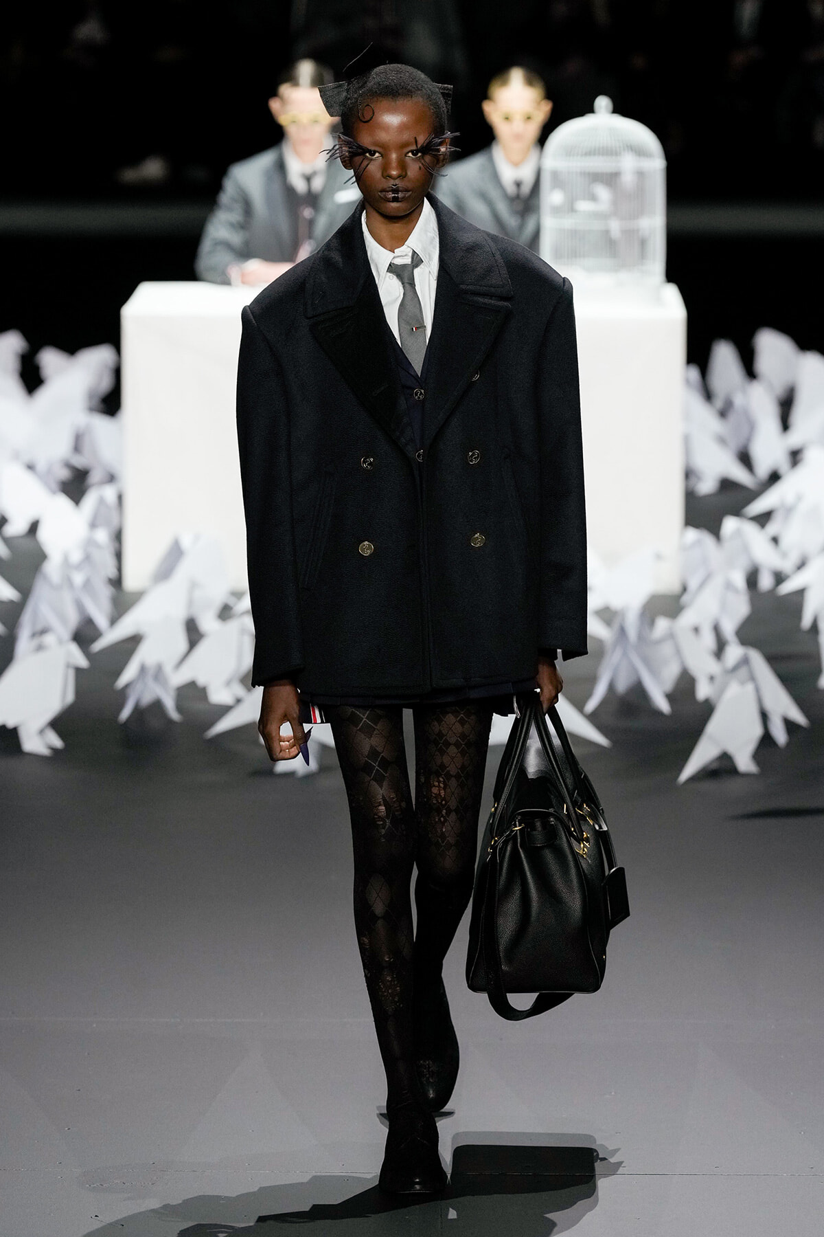 Model in an oversized double-breasted black coat, white shirt, gray tie, patterned tights, and black boots carrying a large black handbag on a fashion runway.
