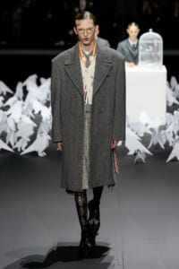 Male model on a runway wearing a oversized gray herringbone overcoat over a layered vest and checkered trousers, with patterned tights and black boots; audience and white paper sculptures in background