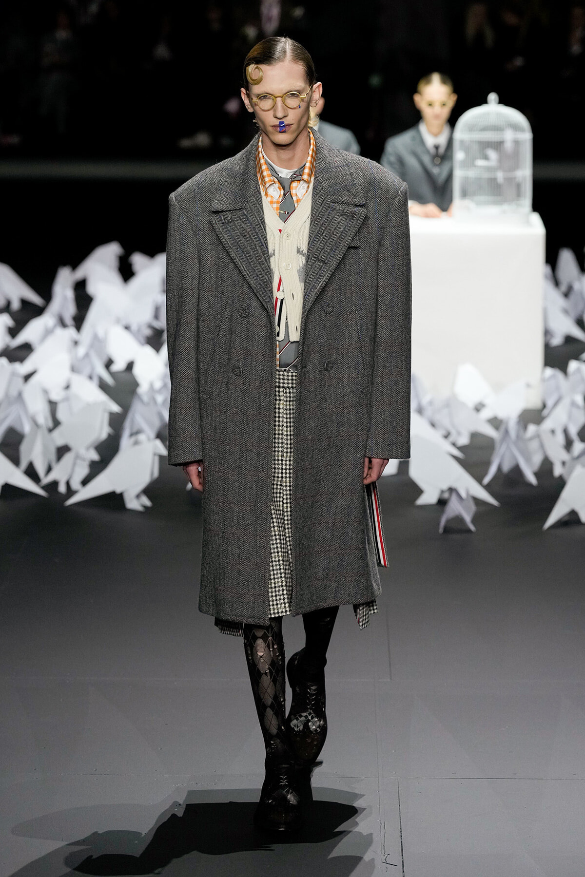 Male model on a runway wearing a oversized gray herringbone overcoat over a layered vest and checkered trousers, with patterned tights and black boots; audience and white paper sculptures in background