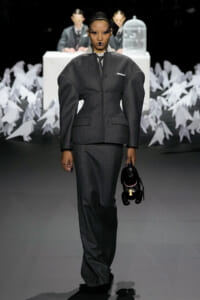 Model on a runway in a tailored gray suit with exaggerated shoulders, holding a small black stuffed dog.