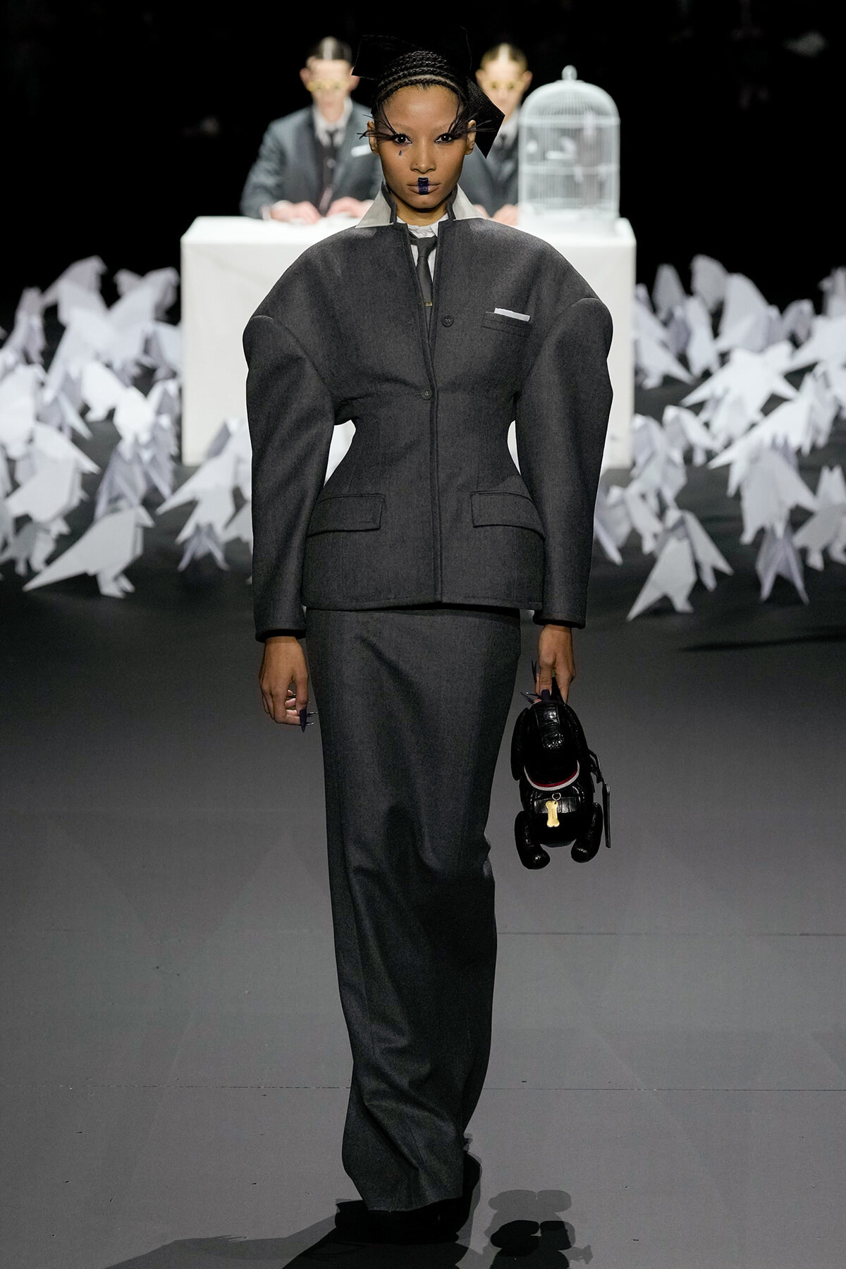 Model on a runway in a tailored gray suit with exaggerated shoulders, holding a small black stuffed dog.