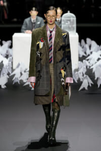 Runway model wearing an oversized patchwork coat with plaid panels, green suede sleeves, and knee-high lace-up boots.