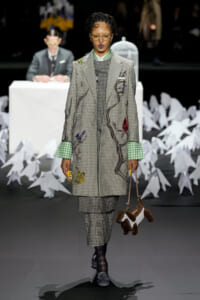 Model walks the runway in a plaid gray suit with embroidered branches and colorful birds, green gingham cuffs, and matching checked trousers, carrying a brown and white animal-shaped handbag.