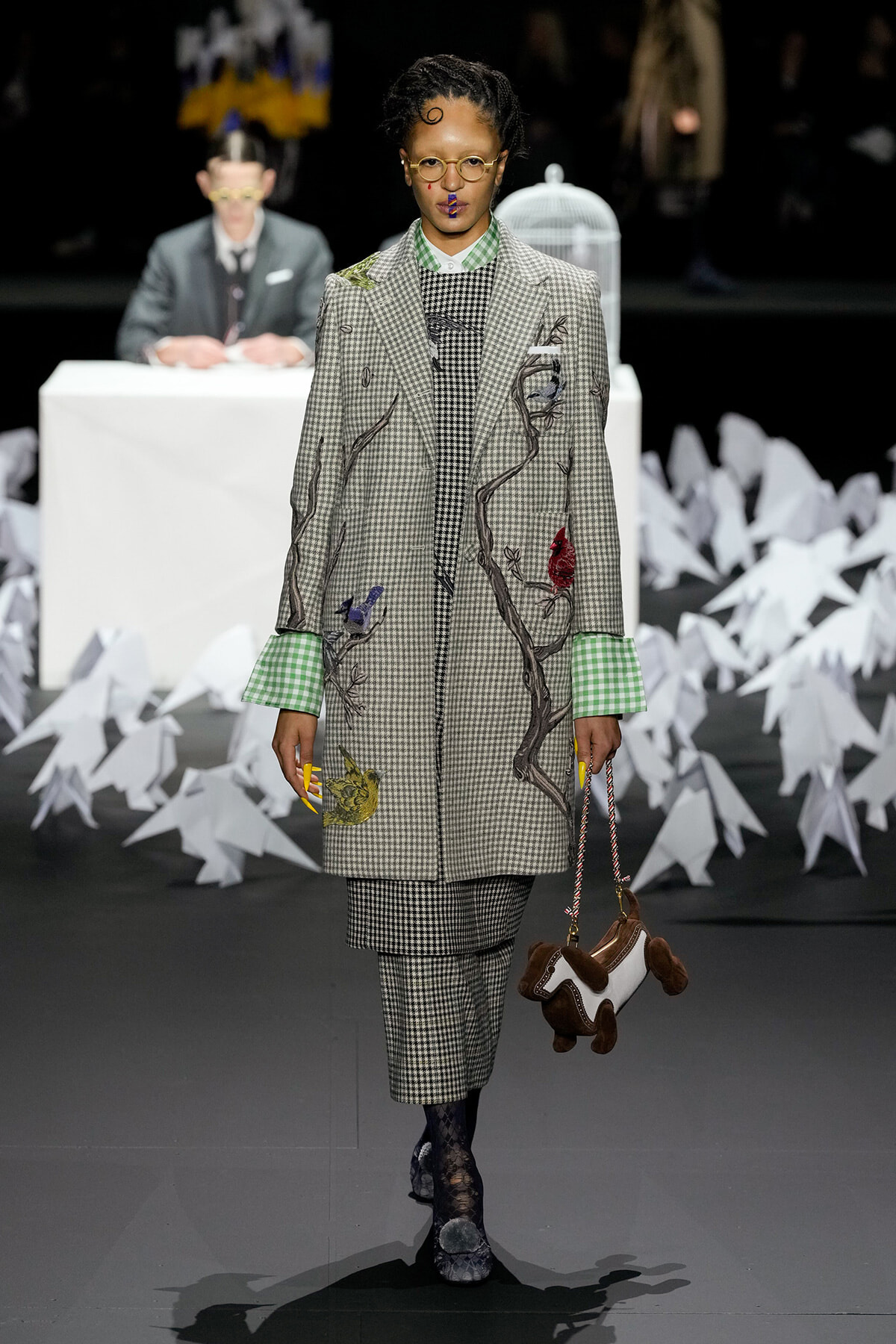 Model walks the runway in a plaid gray suit with embroidered branches and colorful birds, green gingham cuffs, and matching checked trousers, carrying a brown and white animal-shaped handbag.