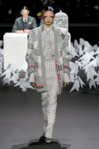 Model walks a fashion runway wearing a chunky gray knit coat over a plaid suit with matching trousers, colorful headpiece, and bold red lashes; geometric paper art backdrop behind.