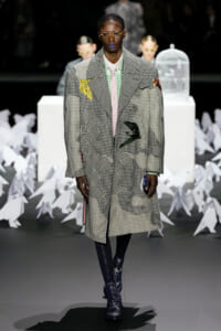 Male model on a runway wearing an oversized patchwork houndstooth coat, pink shirt, green-check collar, round glasses, and blue lipstick.