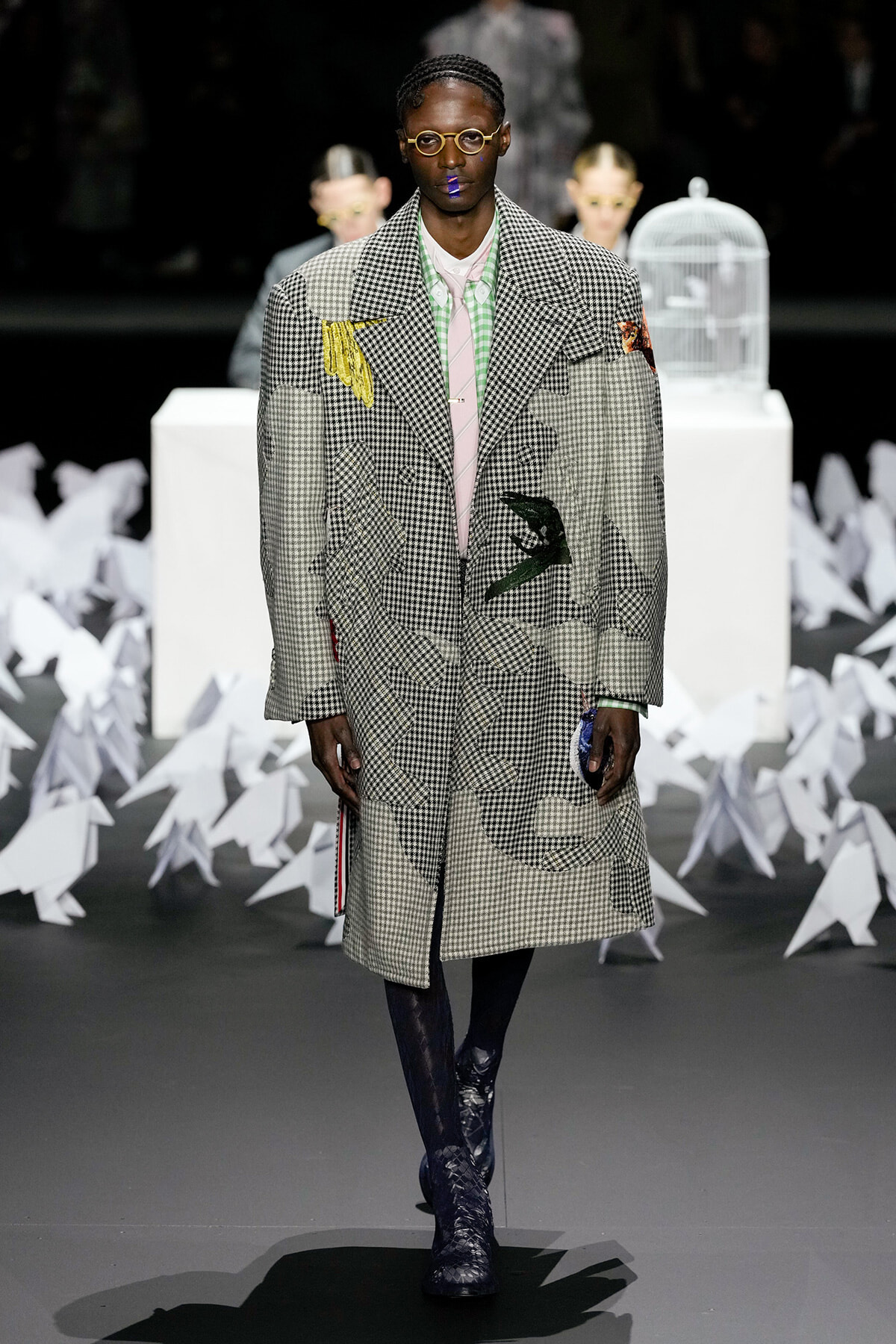 Male model on a runway wearing an oversized patchwork houndstooth coat, pink shirt, green-check collar, round glasses, and blue lipstick.