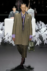 Male model on a runway wearing a brown plaid overcoat, purple gingham shirt, and checkered pants, carrying a green stuffed bunny bag.