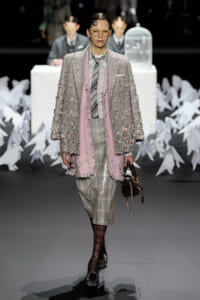 Female model walks the runway in a gray plaid suit with a pink fringe coat and scarf, yellow glasses, and bold lipstick. Behind her are white paper cranes and a white table with a birdcage.