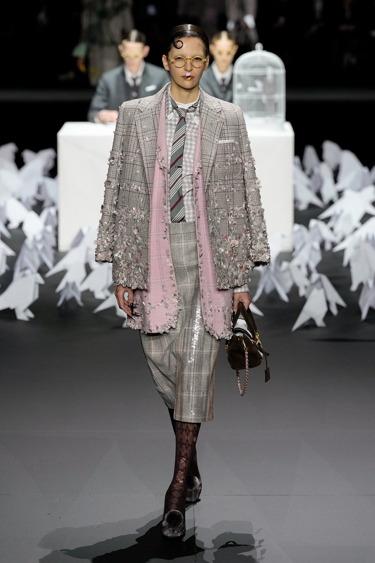 Female model walks the runway in a gray plaid suit with a pink fringe coat and scarf, yellow glasses, and bold lipstick. Behind her are white paper cranes and a white table with a birdcage.