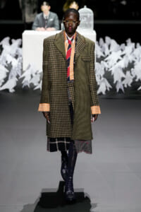 Model walking a runway in a layered oversized plaid coat with orange gingham cuffs and shirt, striped tie, and mismatched plaid skirt pairing.