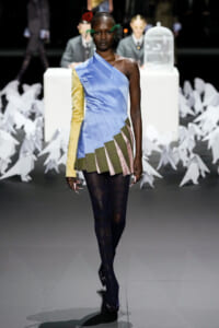 Runway model in a one-shoulder blue dress with a yellow sleeve and layered plaid pleats, paired with dark tights.