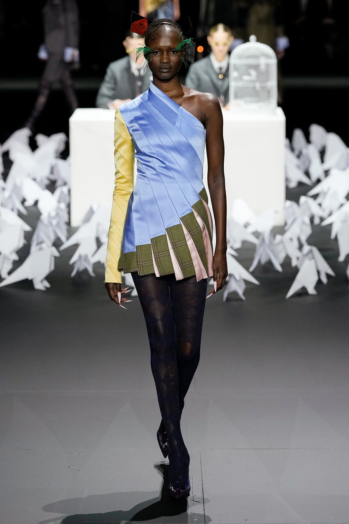 Runway model in a one-shoulder blue dress with a yellow sleeve and layered plaid pleats, paired with dark tights.