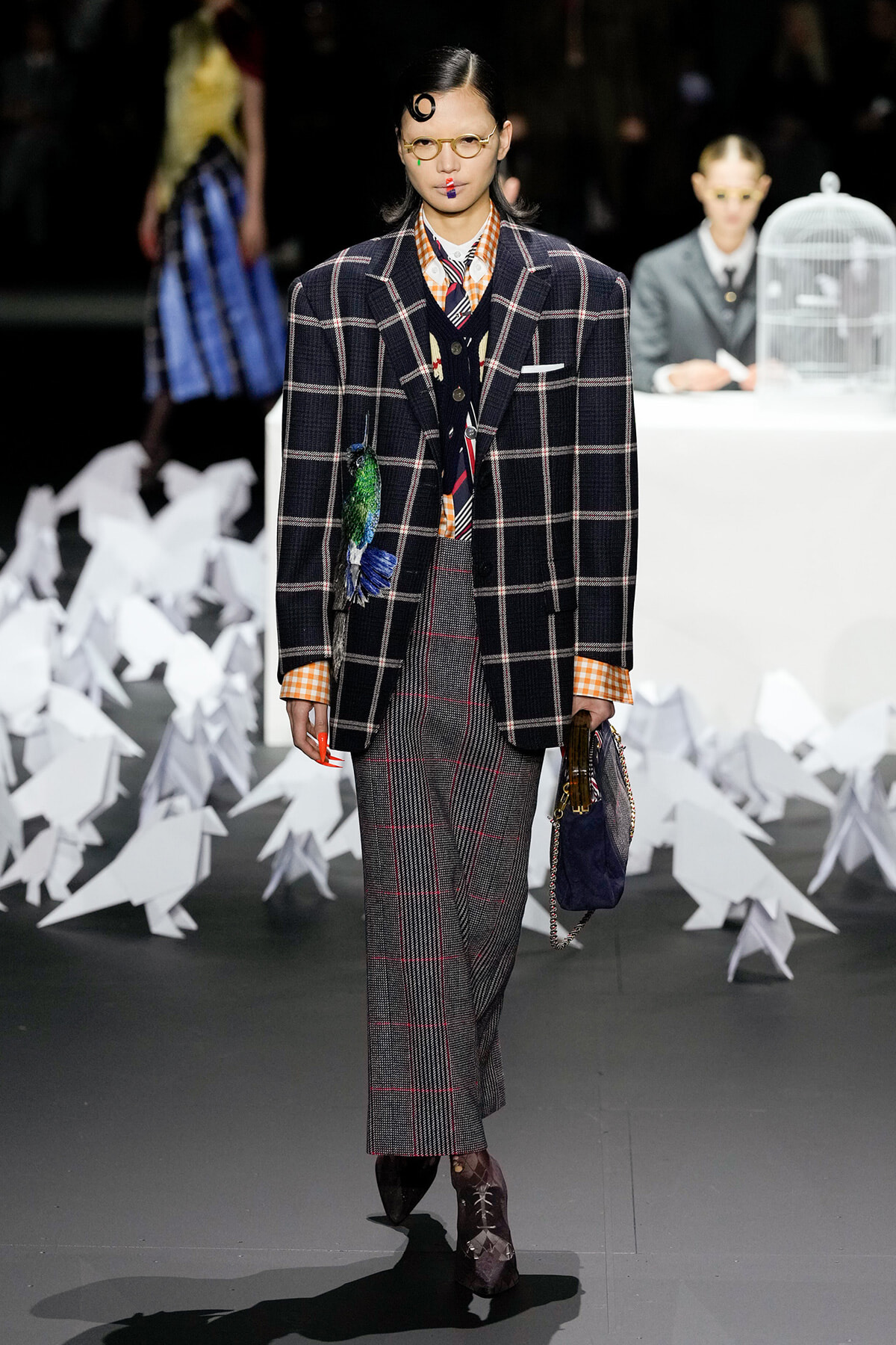 Model walks the runway in a dark plaid oversized blazer over an orange plaid shirt and gray plaid wide-leg pants; yellow round glasses and bold accessories complete the look, with a small handbag in hand. White paper origami shapes line the runway backdrop behind her.