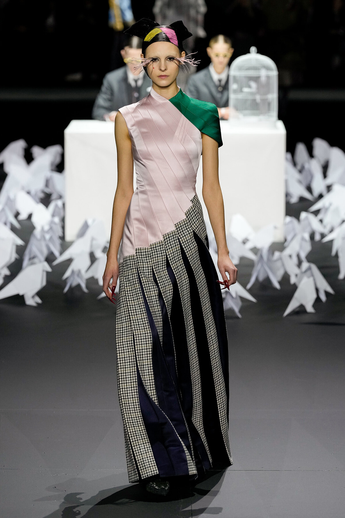 Model walks the runway in a pink asymmetric top with green shoulder, and a plaid-and-navy pleated skirt amid white paper sculptures.