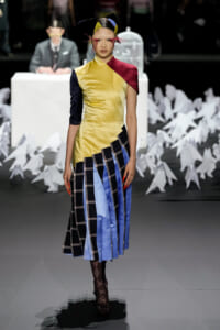 Model on a fashion runway wearing a color-blocked dress with yellow, navy, maroon, and plaid lower panels; black patterned tights and heels.