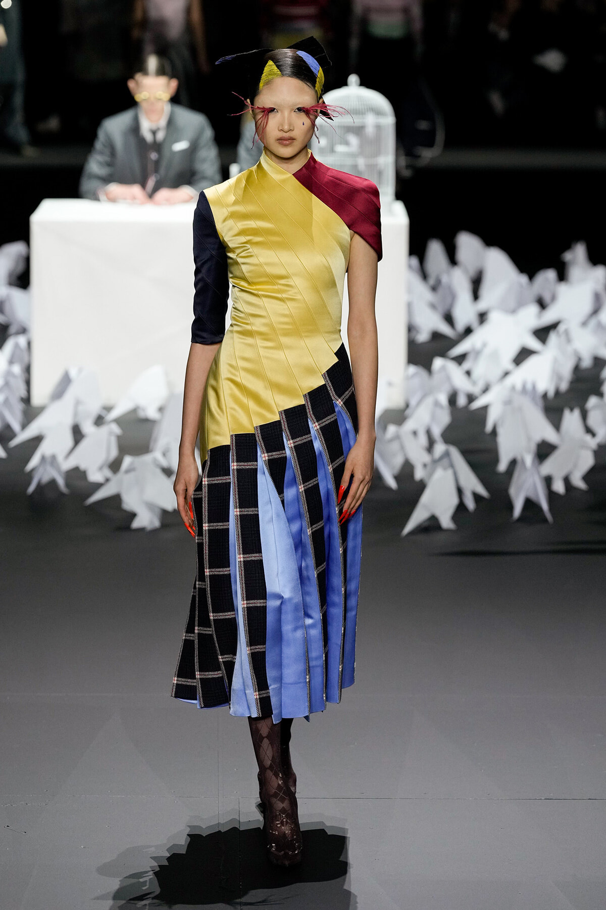 Model on a fashion runway wearing a color-blocked dress with yellow, navy, maroon, and plaid lower panels; black patterned tights and heels.