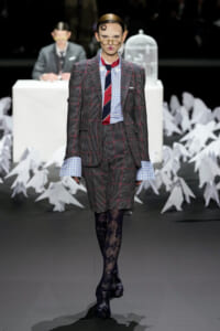 Model on a fashion runway wearing a gray plaid suit with blue gingham shirt cuffs and a red–navy striped tie; white paper birds seen in the background.