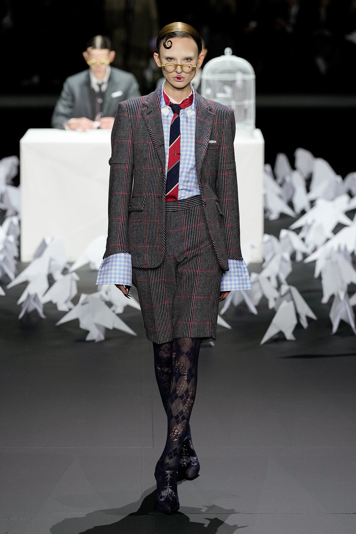 Model on a fashion runway wearing a gray plaid suit with blue gingham shirt cuffs and a red–navy striped tie; white paper birds seen in the background.