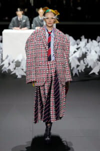 Model on a runway wearing an oversized red-and-white tweed coat over a blue shirt and navy striped tie, with a yellow headpiece and white feather accents.