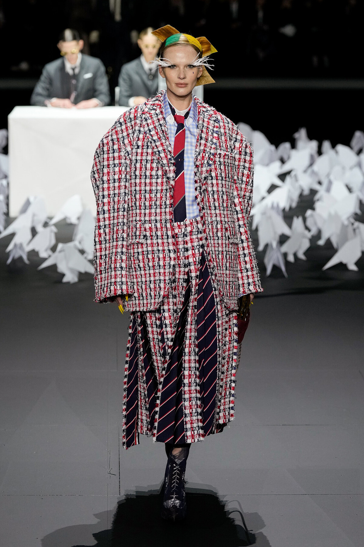 Model on a runway wearing an oversized red-and-white tweed coat over a blue shirt and navy striped tie, with a yellow headpiece and white feather accents.