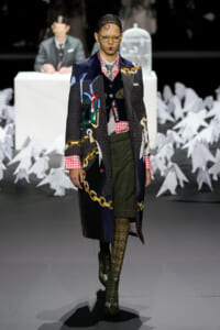 Model walking a runway in a long patchwork coat with gold chain patterns, over a red plaid shirt, green plaid skirt, and patterned tights with knee-high boots; a white birdcage prop and judge table are in the blurred background.