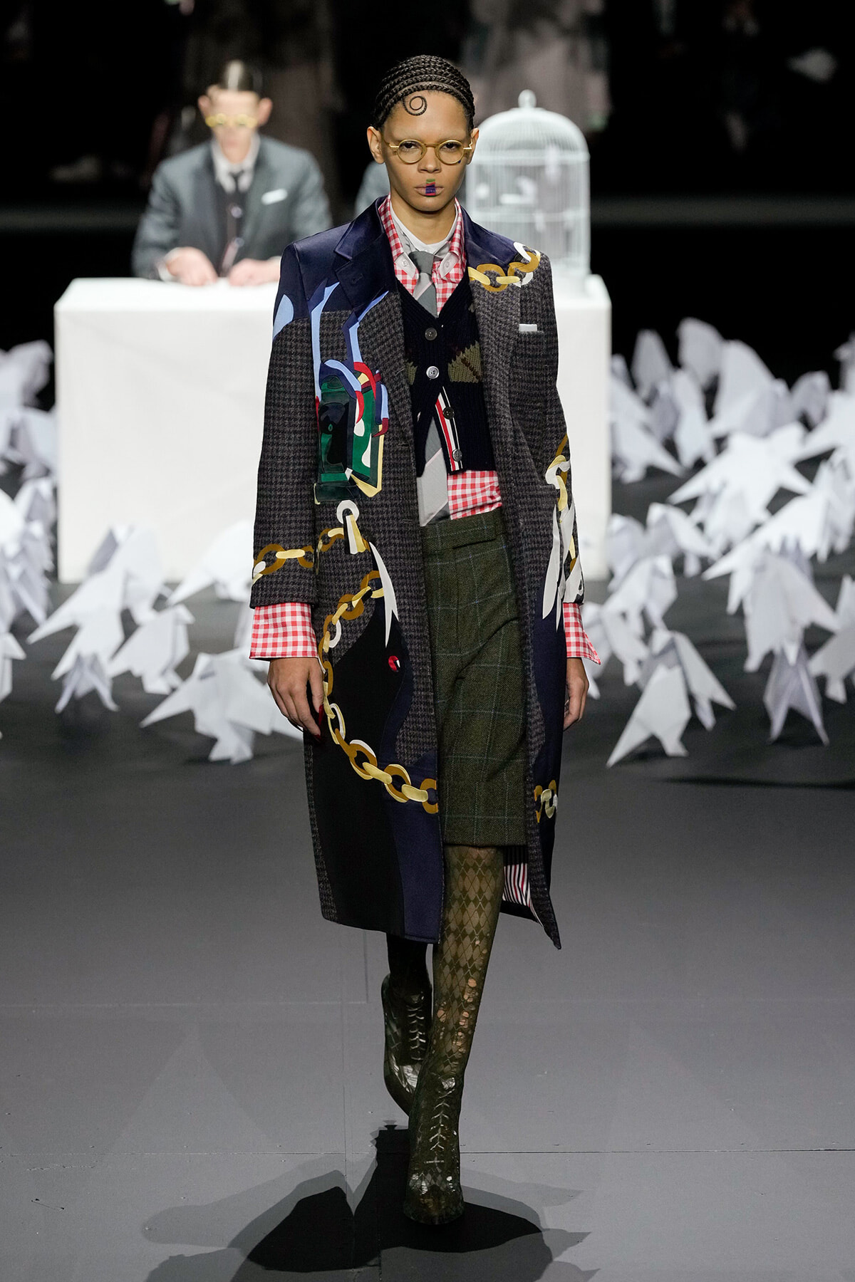 Model walking a runway in a long patchwork coat with gold chain patterns, over a red plaid shirt, green plaid skirt, and patterned tights with knee-high boots; a white birdcage prop and judge table are in the blurred background.