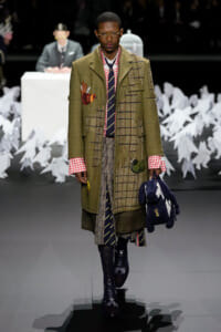 Male model in olive green patched overcoat with red gingham cuffs, plaid skirt layers, striped tie, and round glasses walking a runway. He carries a dark blue plush bag.