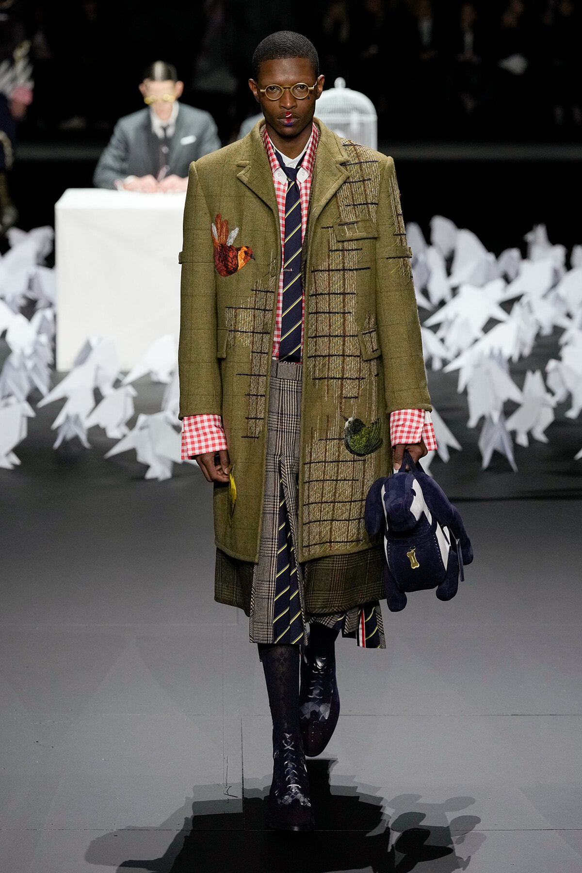 Male model in olive green patched overcoat with red gingham cuffs, plaid skirt layers, striped tie, and round glasses walking a runway. He carries a dark blue plush bag.