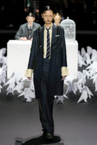 Model on a runway wearing a dark oversized suit with black bead embellishments, yellow gingham shirt, and striped tie; round glasses and bold makeup.