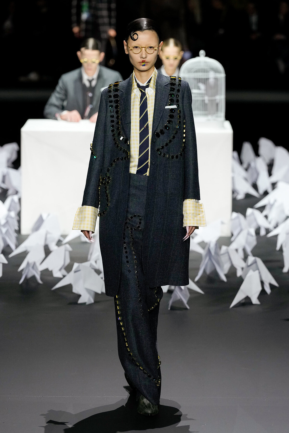 Model on a runway wearing a dark oversized suit with black bead embellishments, yellow gingham shirt, and striped tie; round glasses and bold makeup.