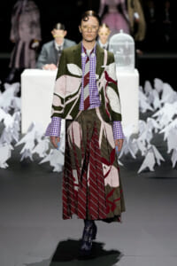 Model on a fashion runway wearing a bold, patchwork suit with green, pink, and maroon panels, a purple checkered shirt, and a tie; pleated skirt and lace-up boots, with paper cranes lining the backdrop.