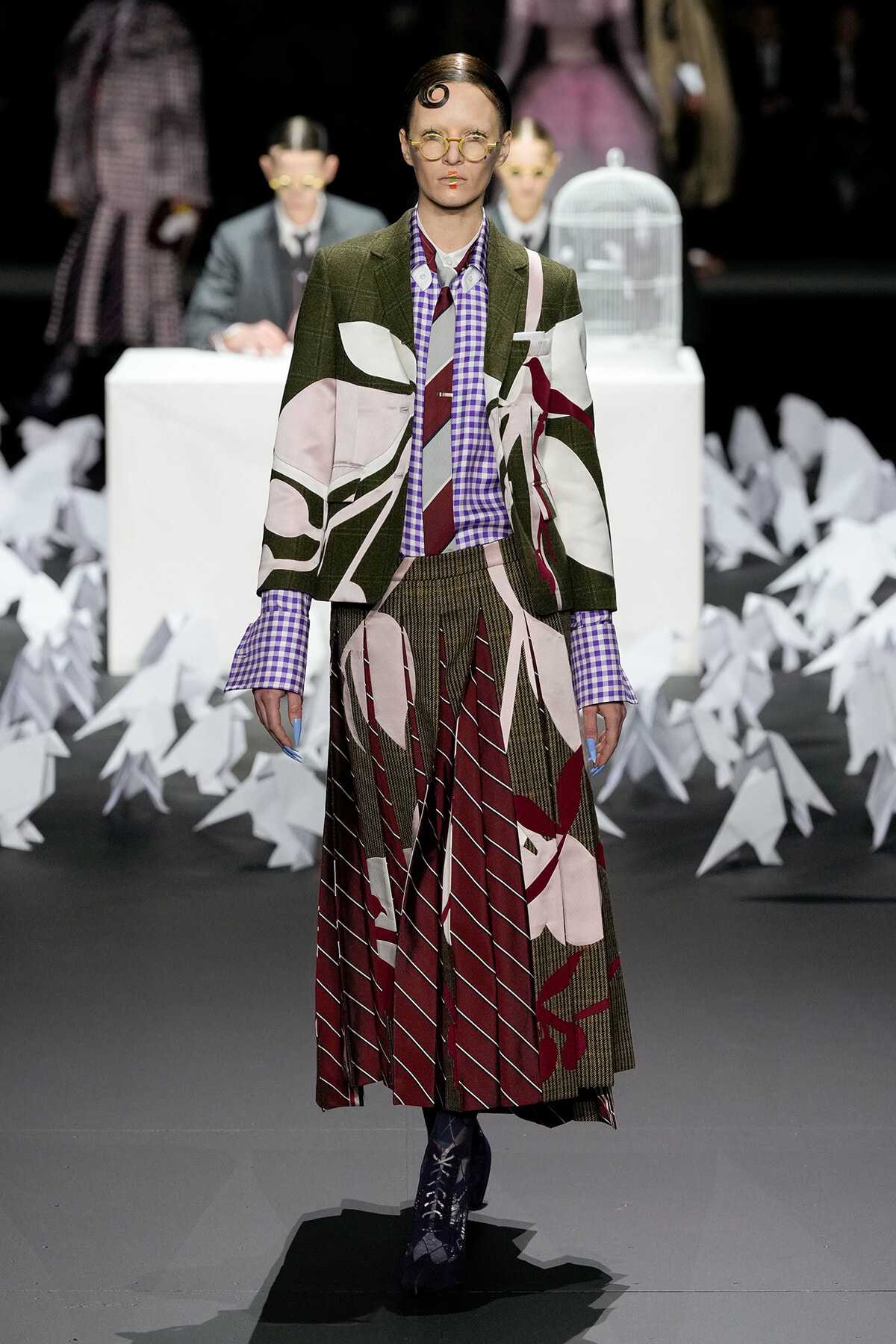 Model on a fashion runway wearing a bold, patchwork suit with green, pink, and maroon panels, a purple checkered shirt, and a tie; pleated skirt and lace-up boots, with paper cranes lining the backdrop.