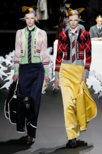 Two models walk a fashion runway in eclectic knitwear: pastel pink and green argyle cardigans over gingham shirts, long dark navy and bright yellow satin skirts, and bold headpieces with abstract shapes.