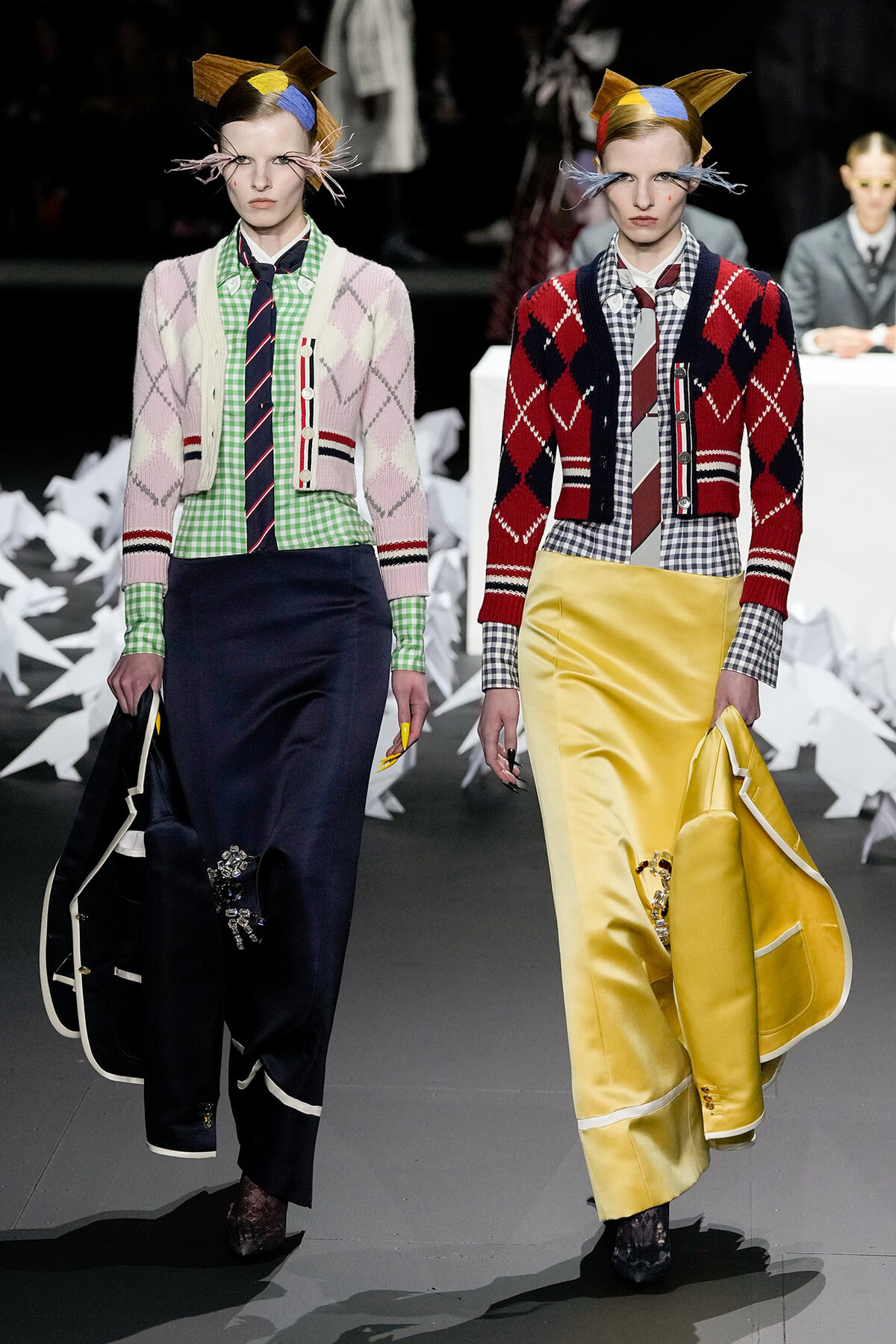 Two models walk a fashion runway in eclectic knitwear: pastel pink and green argyle cardigans over gingham shirts, long dark navy and bright yellow satin skirts, and bold headpieces with abstract shapes.