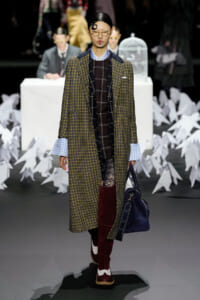Model walks a fashion show wearing a long yellow‑black plaid overcoat over a brown plaid dress, blue‑check shirt cuffs, burgundy knee‑high boots, round glasses, and a navy bag.