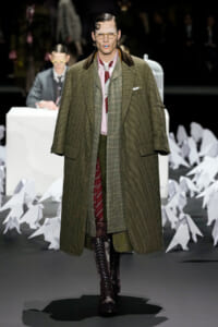 Male model on a fashion runway wearing a oversized green-brown checkered coat over layered suit and burgundy knee-high lace-up boots.