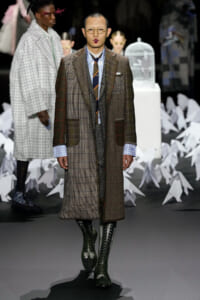 Male model on a fashion runway wearing a brown plaid overcoat with a blue check shirt, orange striped tie, and tall lace-up boots; round glasses.