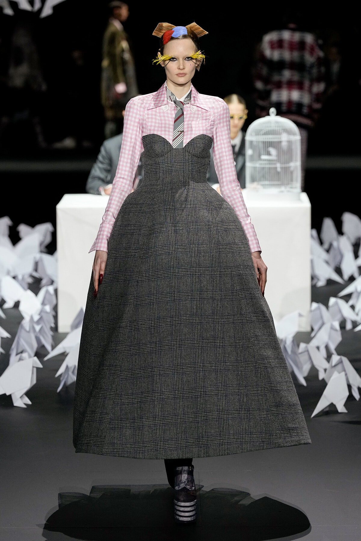 Model walks the runway in a gray plaid, full-skirted gown paired with a pink gingham blouse, striped necktie, and a multicolored bow headpiece; bold yellow feathered eye accents add whimsy to the look.