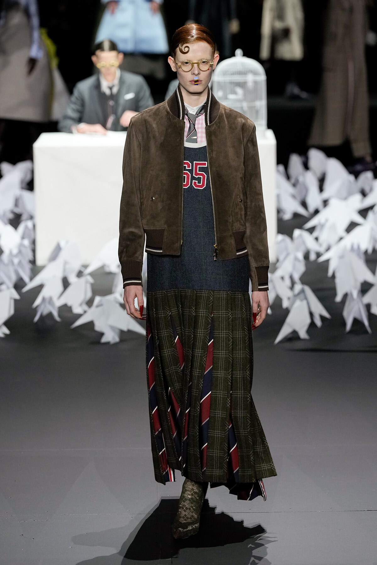 Model walks the runway in a brown suede varsity-style jacket over a layered dress, with round glasses and a quirky curl in the hair.