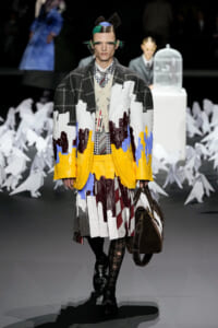 Model walking a fashion runway wearing a oversized patchwork coat in yellow, white, gray, and blue with a plaid shirt and striped tie, carrying a brown bag.