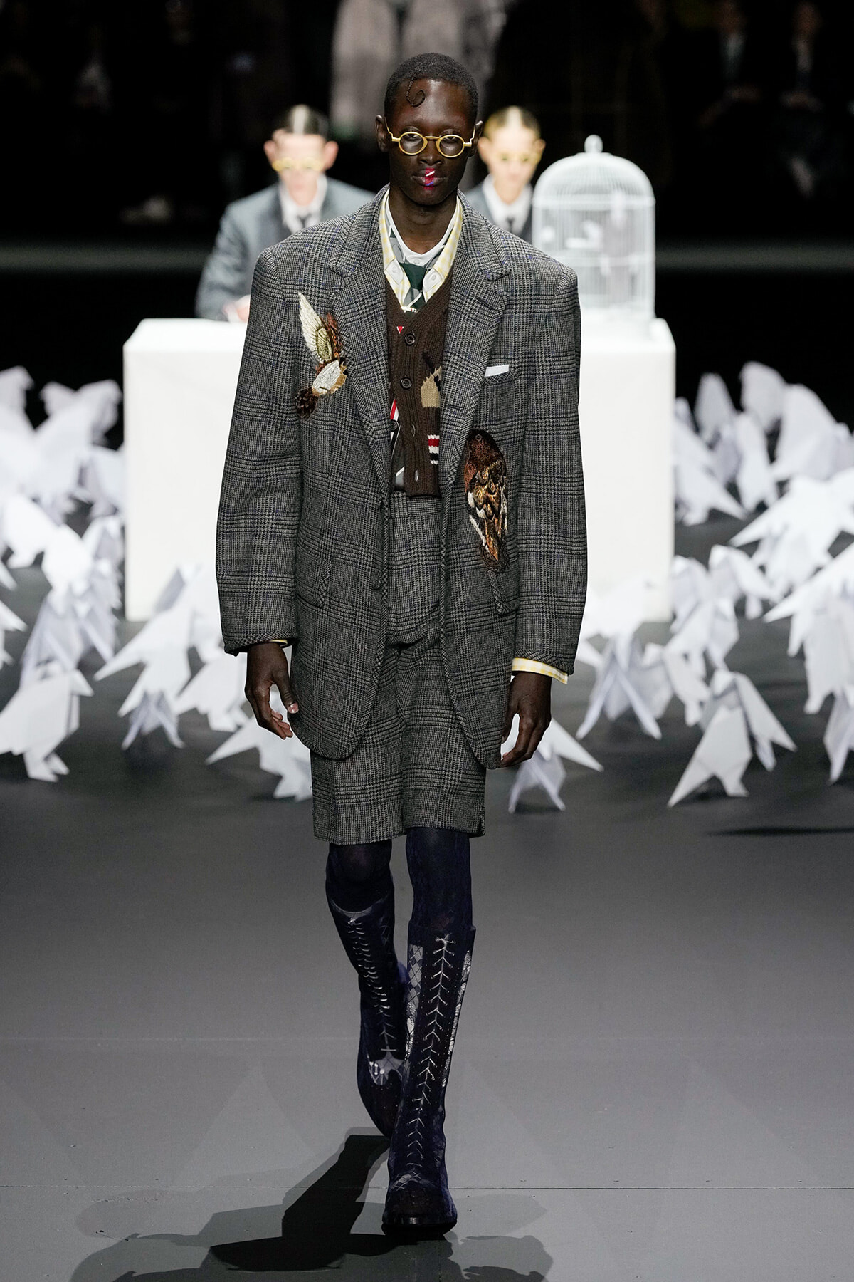 Male model walking a fashion runway in a gray plaid suit with large embroidery patches, wearing knee-high blue boots and round glasses.
