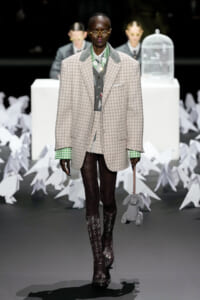 Runway model in an oversized beige checkered blazer with green cuffs, gray vest, and tights, holding a gray stuffed rabbit on a chain.