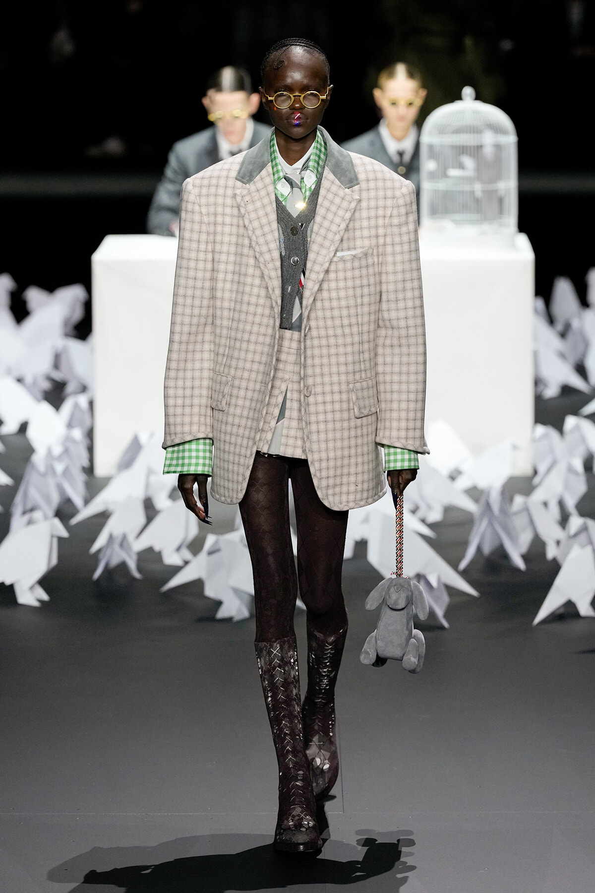 Runway model in an oversized beige checkered blazer with green cuffs, gray vest, and tights, holding a gray stuffed rabbit on a chain.