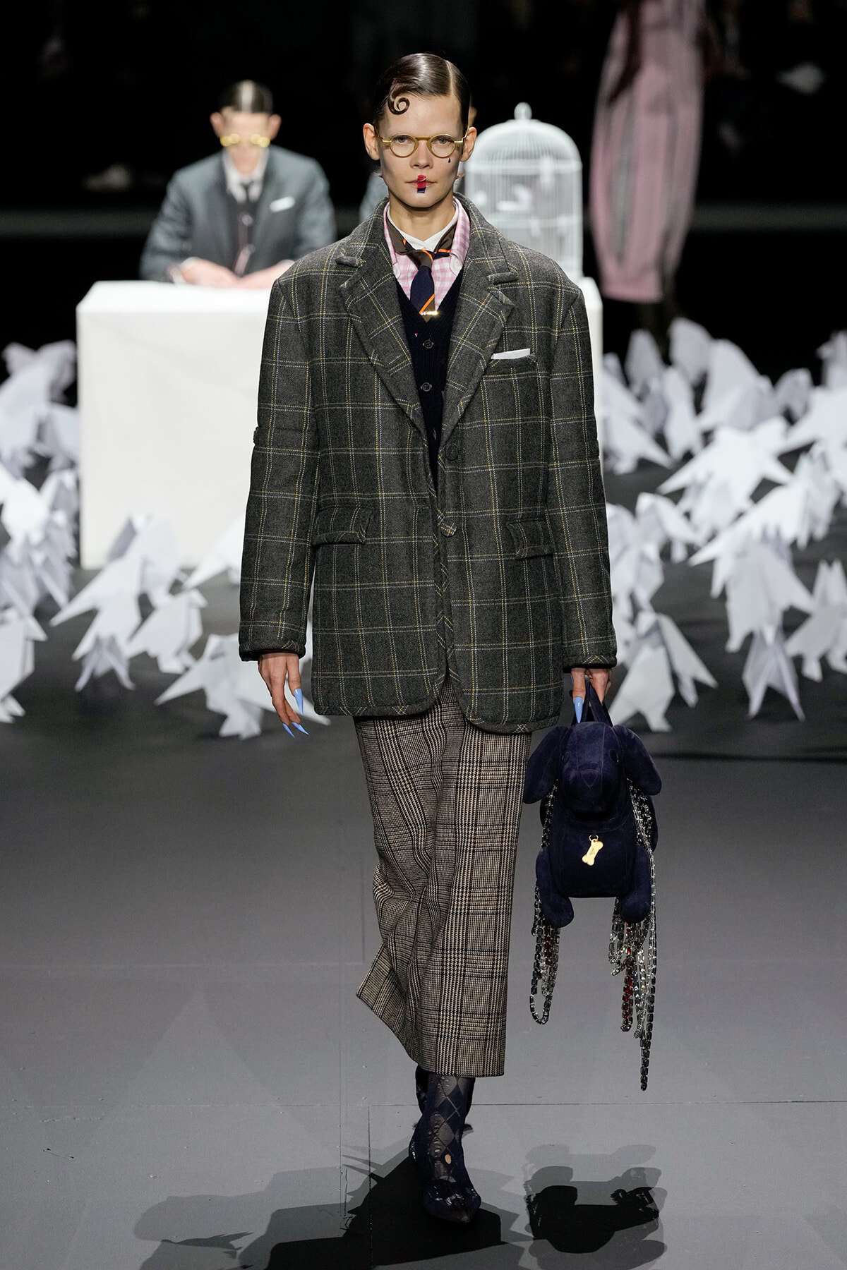 Model on a fashion runway wearing an oversized dark plaid blazer with matching checkered trousers, pink shirt, and round glasses, carrying a navy plush backpack with chain tassels.