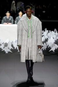 Male runway model in a heavy gray plaid coat with a matching skirt, green check shirt, black patterned tights, and a gray handbag; white decor in the background.