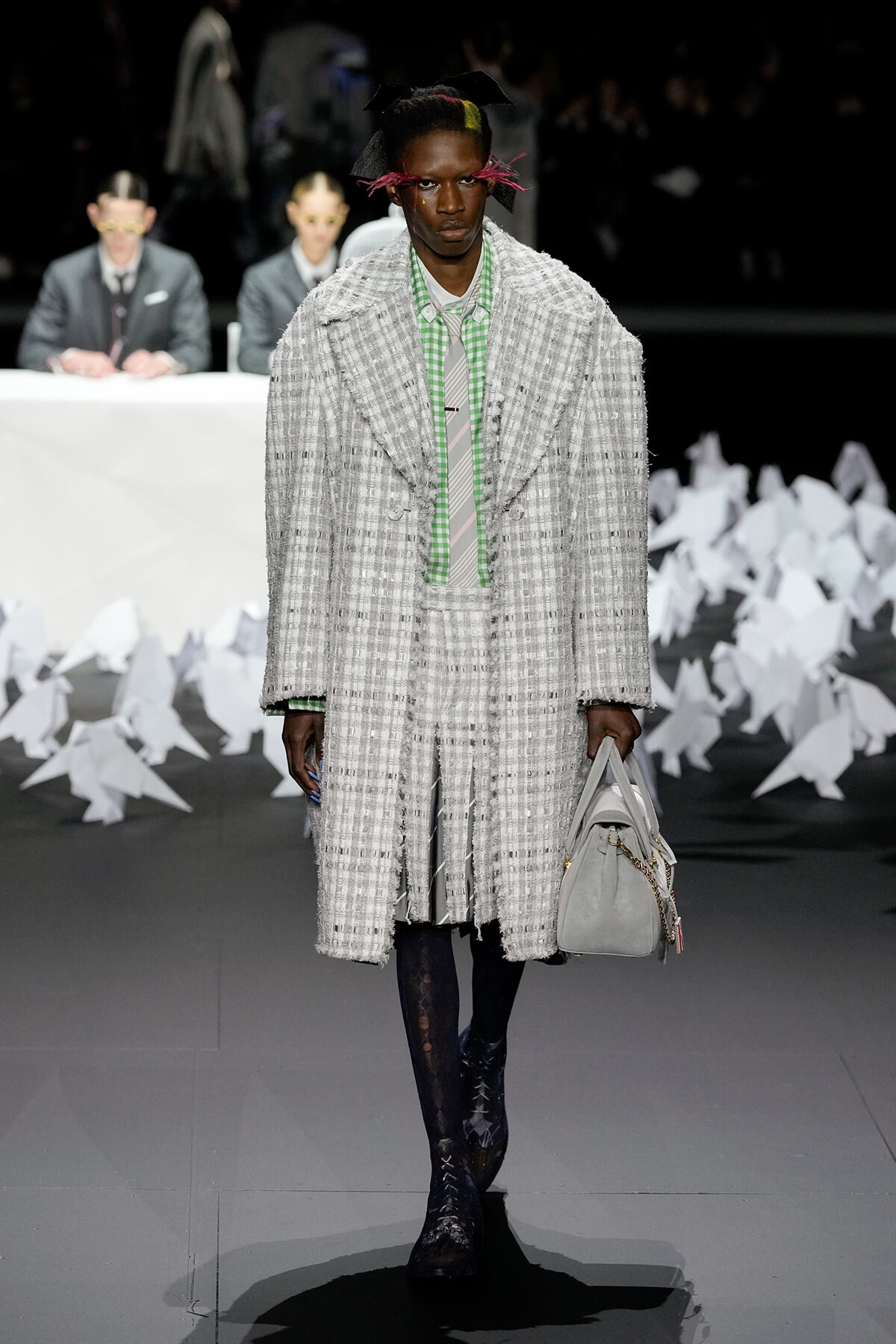 Male runway model in a heavy gray plaid coat with a matching skirt, green check shirt, black patterned tights, and a gray handbag; white decor in the background.