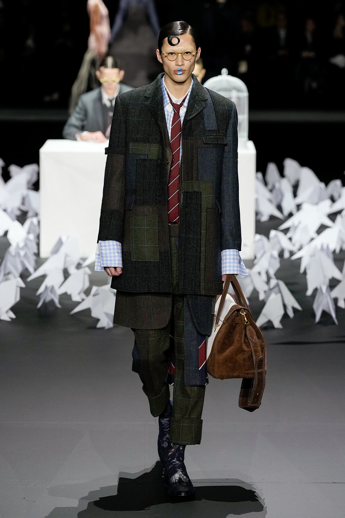 Male model walks the runway in a dark patchwork suit with a burgundy striped tie and blue-check shirt, carrying a brown leather duffel bag.