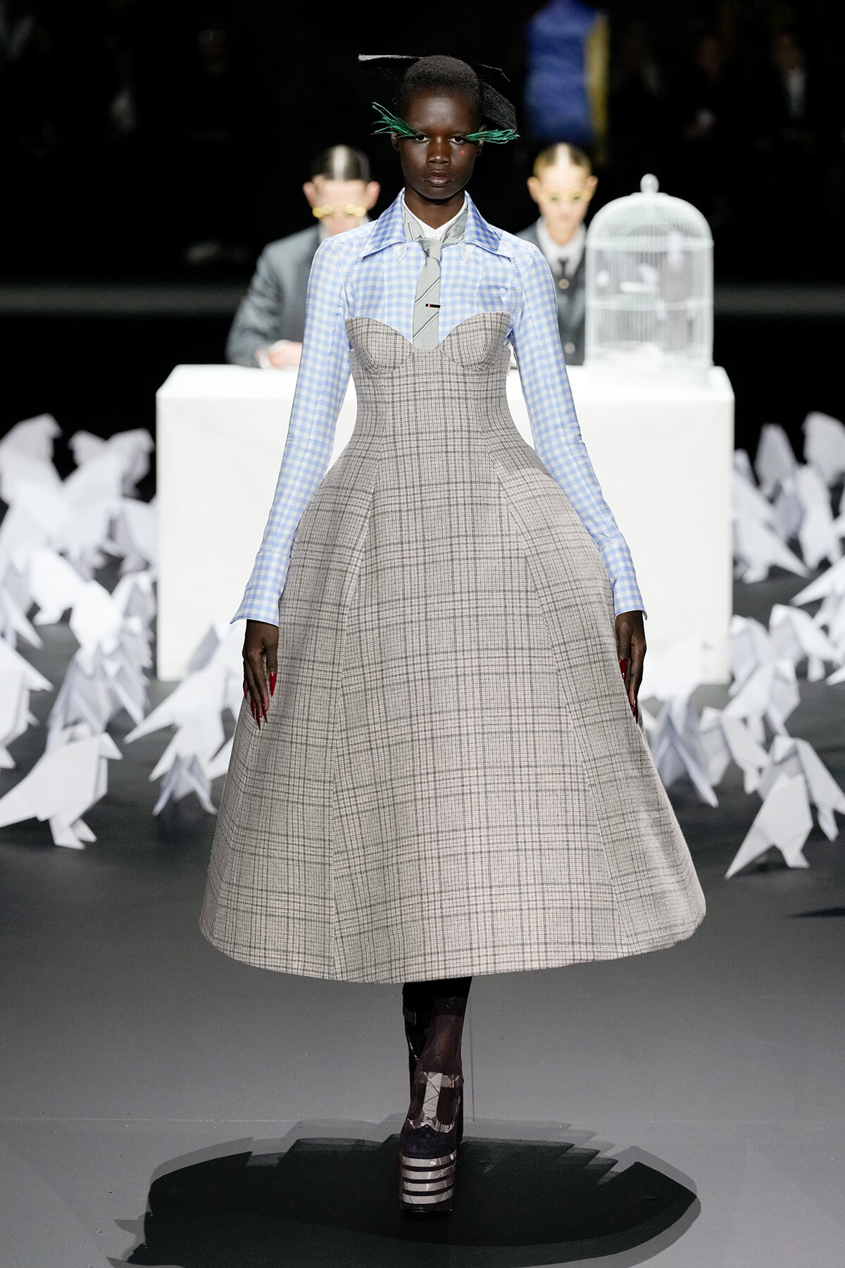 Model walks the runway in a gray plaid A-line dress layered over a light blue gingham shirt with a gray tie, black platform heels visible.