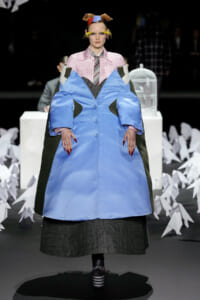Runway model in an oversized light-blue coat with dark panels, wearing a pink checkered shirt and striped tie, among white paper crane props.